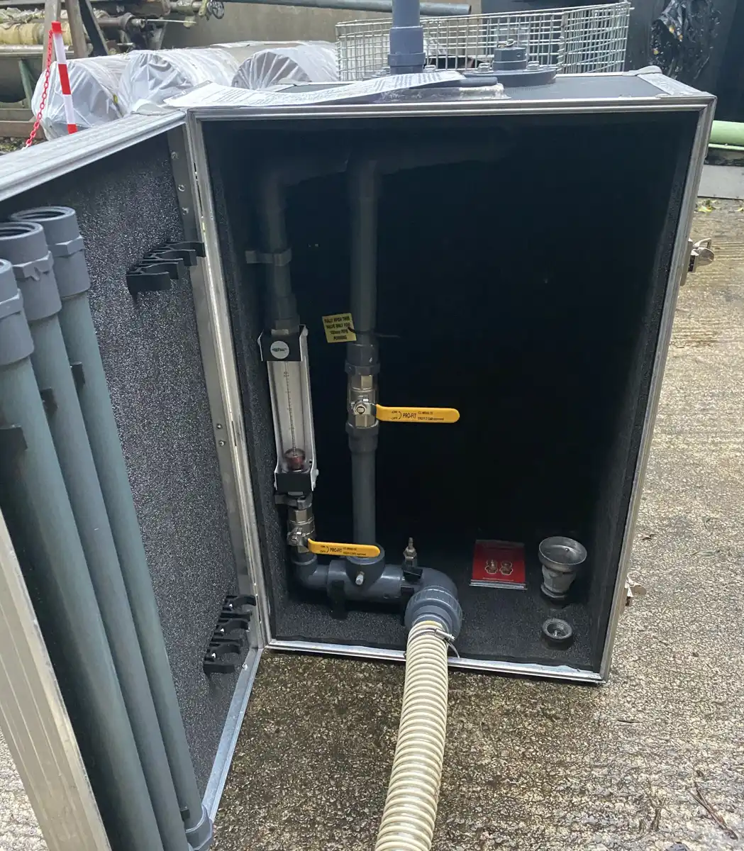 Open flight case holding a portable pipework rig with a flow meter, two yellow-handled valves and a flexible hose