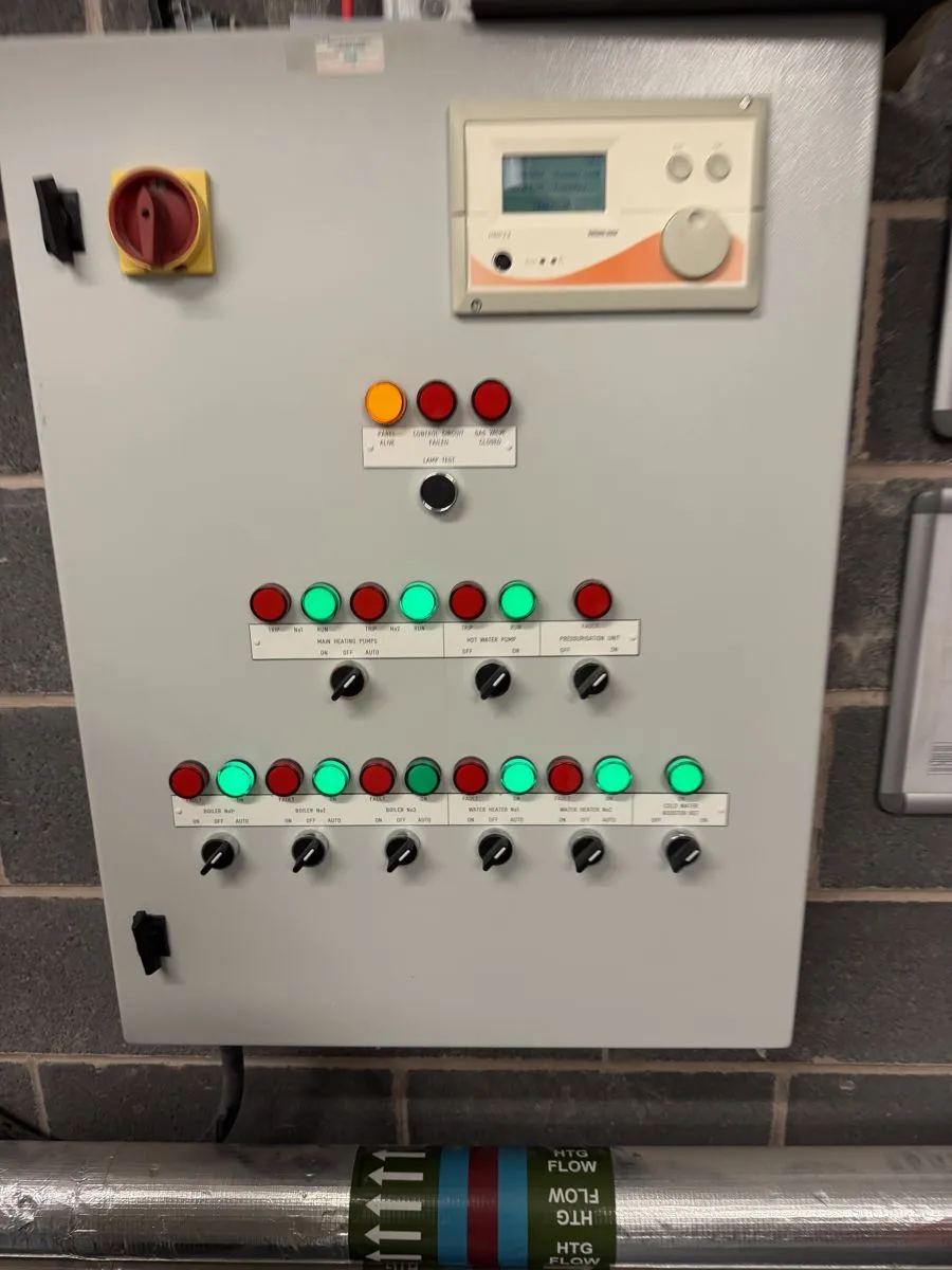 Boiler plant control panel showing boiler, heating pump and hot water pump status lights and selector switches, above insulated heating flow pipework