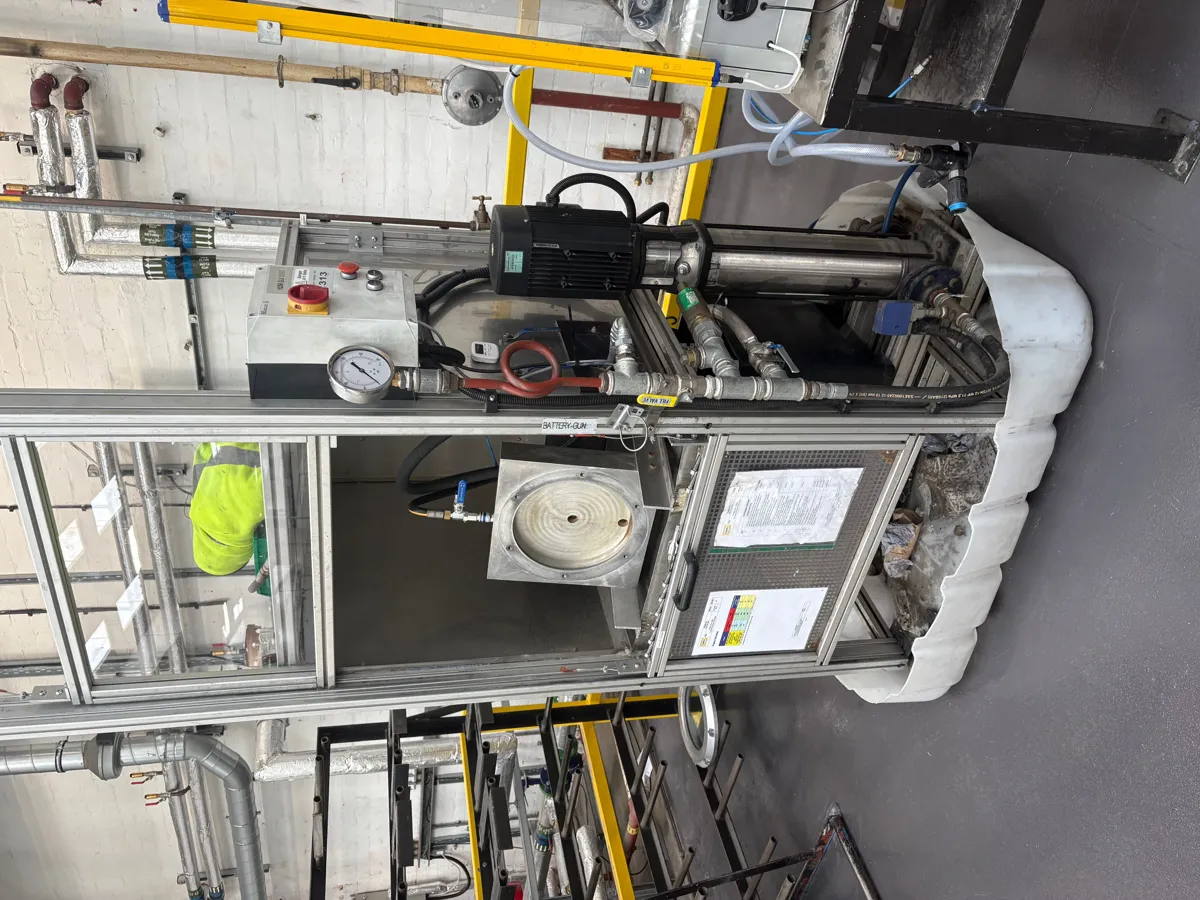 Aluminium-framed pump rig with a vertical pump, pressure gauge and control box standing on a drip tray in a workshop