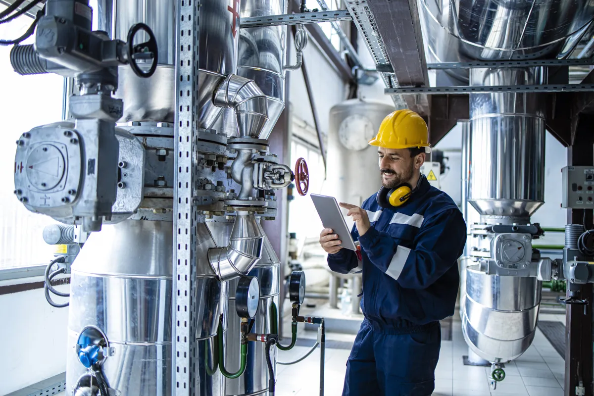 Engineer in a yellow hard hat checking a tablet beside insulated pipework and valves in a plant room